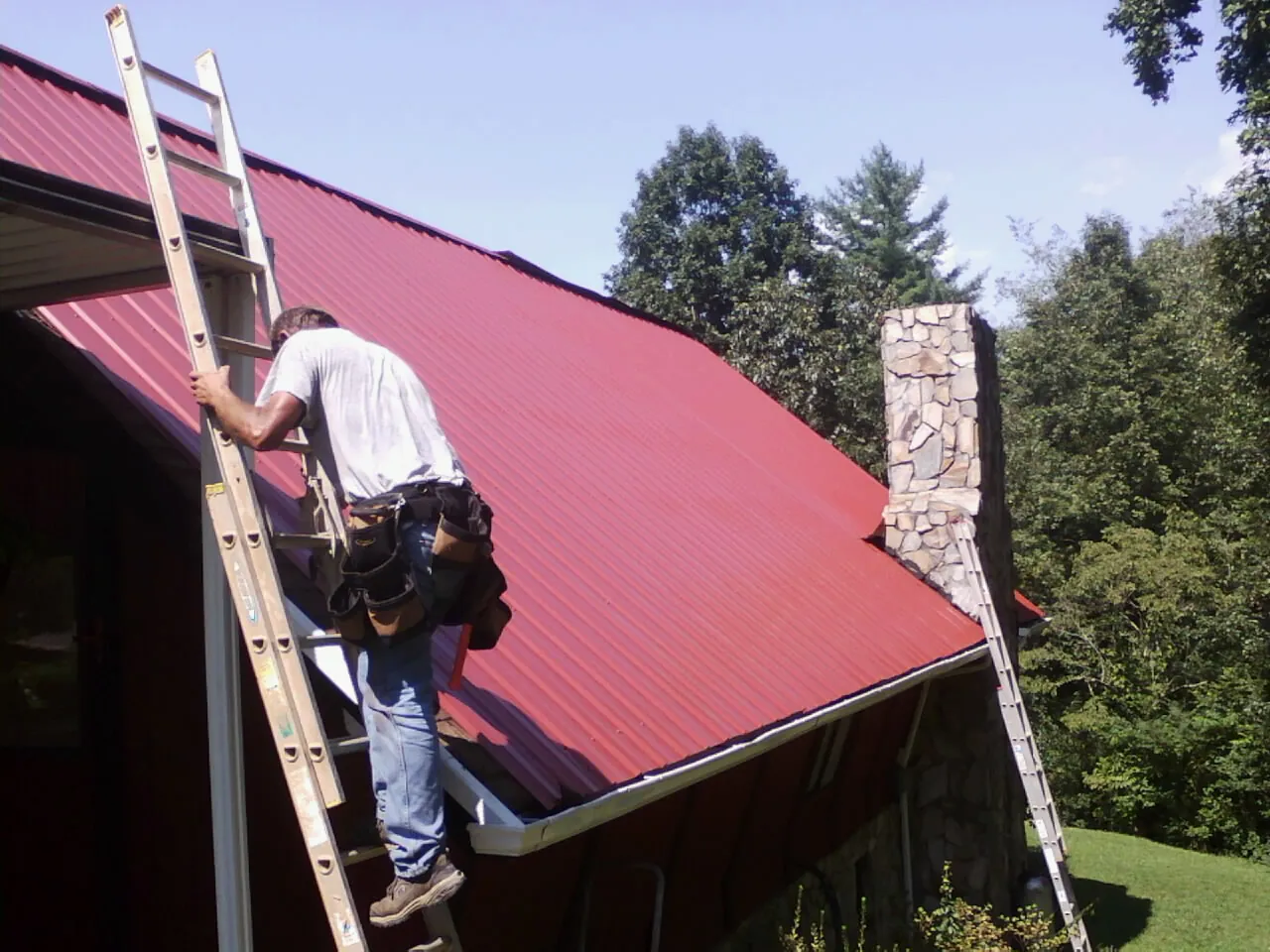 Roofer on a ladder beside a red metal roof and stone chimney — Soffit & Fascia Repair by Roofing Brothers in Graysville